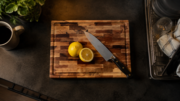 Hard maple cutting board on a small apartment kitchen counter with a chef's knife and halved lemon, mug and dish rack just in frame