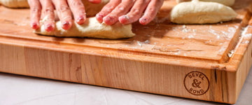 Hands shaping bread dough on a thick end-grain maple block, logo on the side edge