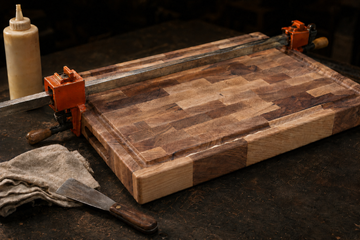End-grain cutting board in bar clamps during a glue joint repair, with wood glue bottle and putty knife on a dark workbench