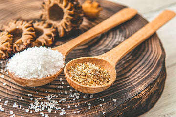 Two wooden spoons holding coarse salt and ground pepper on a dark wood slab